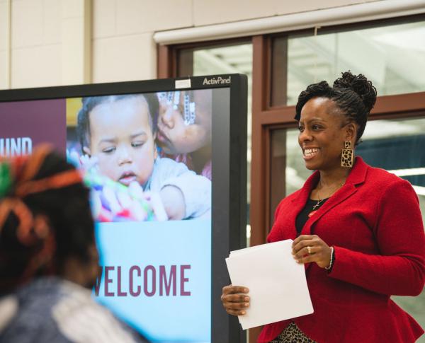 A Black woman wearing a red blazer and leopard pants holds a piece of paper in front of a presentation screen, leading a class of adult families.