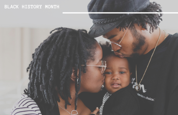 A Black mother and father kiss a young baby.