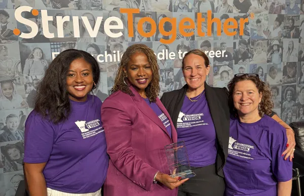 Four women stand with their arms around each other with one holding an award. All are wearing purple tops.