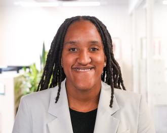A young Black woman with locs smiles at the camera, wearing a black shirt and light mint blazer.