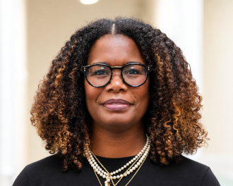 A young Black woman with natural hair and multiple necklaces wearing a black shirt smiles at the camera