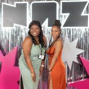 Two women in formal dresses smile for the camera in front of a large block lettering reading "NAZ"