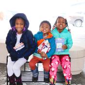 Three young children sit in their winter jackets holding books