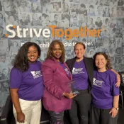 Four women stand with their arms around each other with one holding an award. All are wearing purple tops.