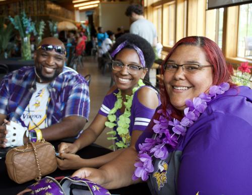 Three people sit in a row, all wearing flowers around their neck.