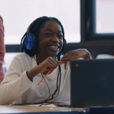 Young girl smiling, wearing headphones, sitting behind a computer