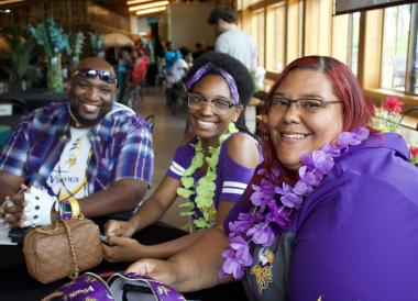 Three people sit in a row, all wearing flowers around their neck.