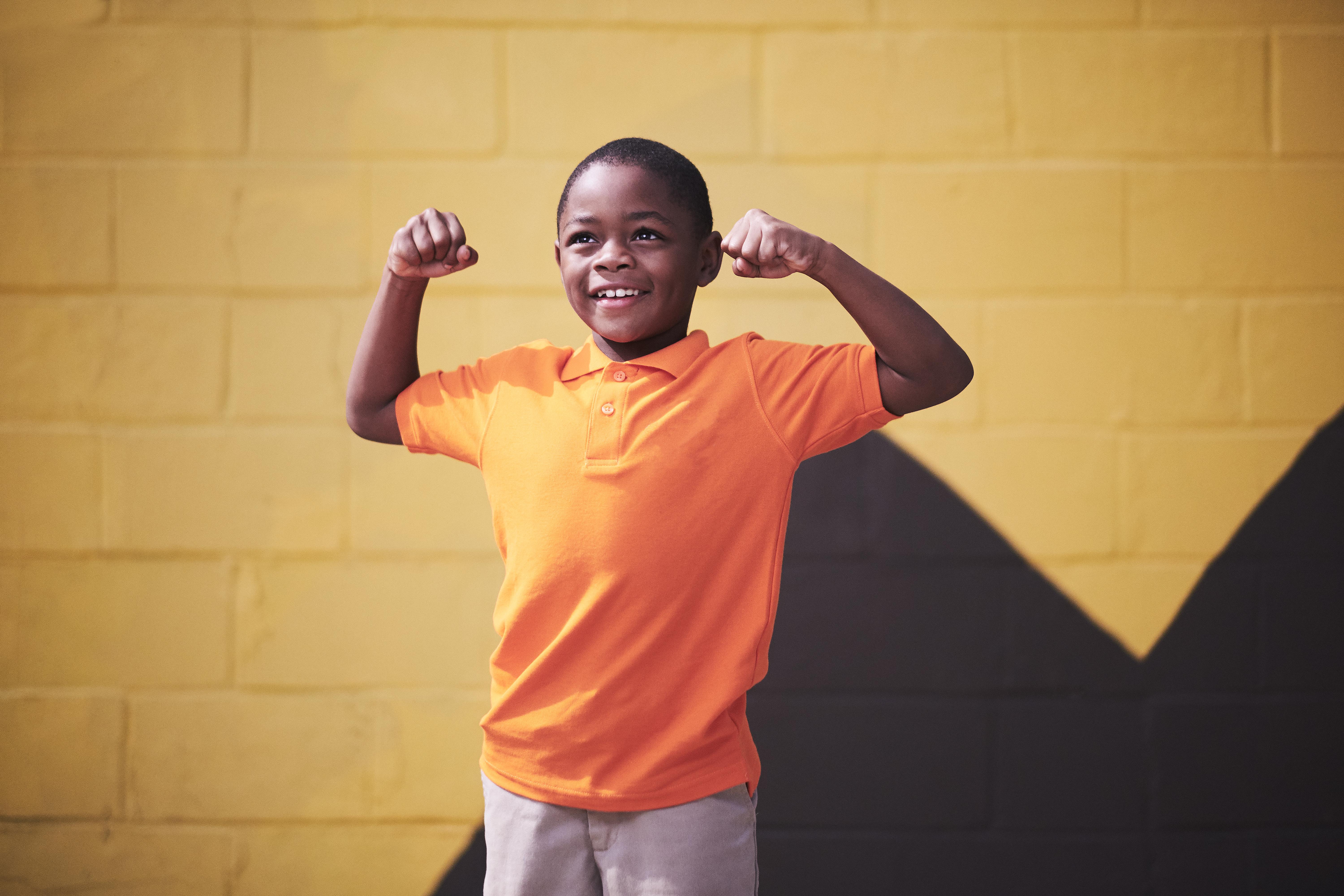 A young Black boy wears an orange shirt and smiles, flexing his arms.