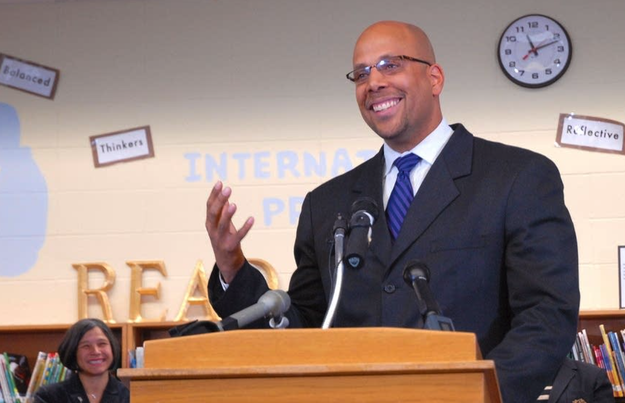 Jim Shelton stands at a podium, addressing a crowd of people, in a library.