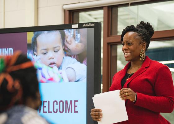 A Black woman wearing a red blazer and leopard pants holds a piece of paper in front of a presentation screen, leading a class of adult families.