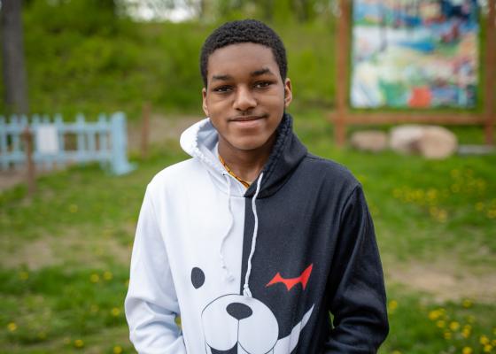 A young Black man stands in front of a hill, wearing a black and white hoodie. He's smiling.