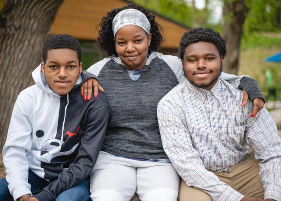A Black mother sits with her two high school aged sons. They're outside, under two trees.