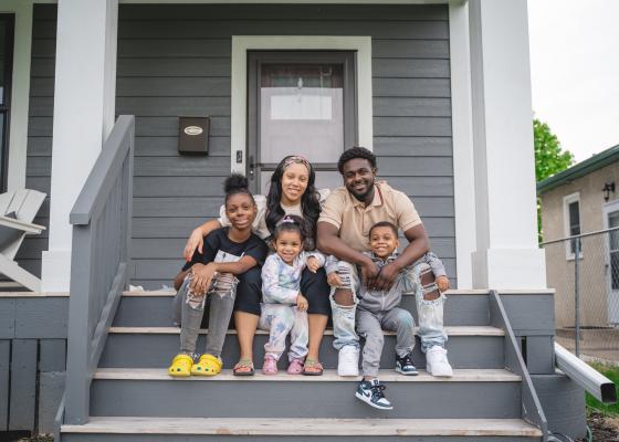 A mother, father and their two daughters and son sit outside their house on the front porch. They're all smiling.