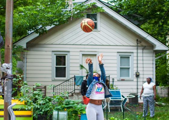 A young high school girl plays basketball in the backyard. The net is frayed and the ball hasn't gone through yet.