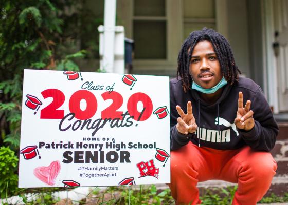 A young Black man, who is a senior in high school, squats next to a yard sign that says "Class of 2020, Congrats!"