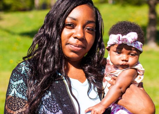 A young Black mother wears a lace sweater and blue shirt, holding a baby girl, who is 2 months old.