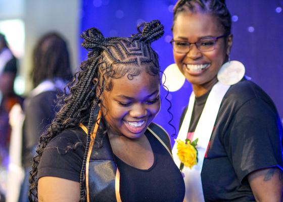 Young woman wearing a graduation stole with a woman behind her smiling