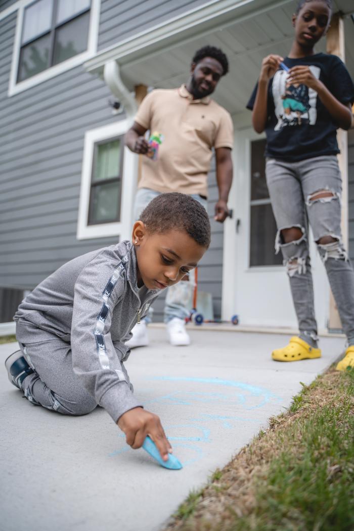 Young boy plays with chalk in their family's backyard. Sister and dad look over.