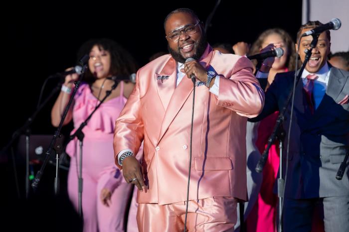 A chorus director stands in front of a smiling group of young people, microphone in hand