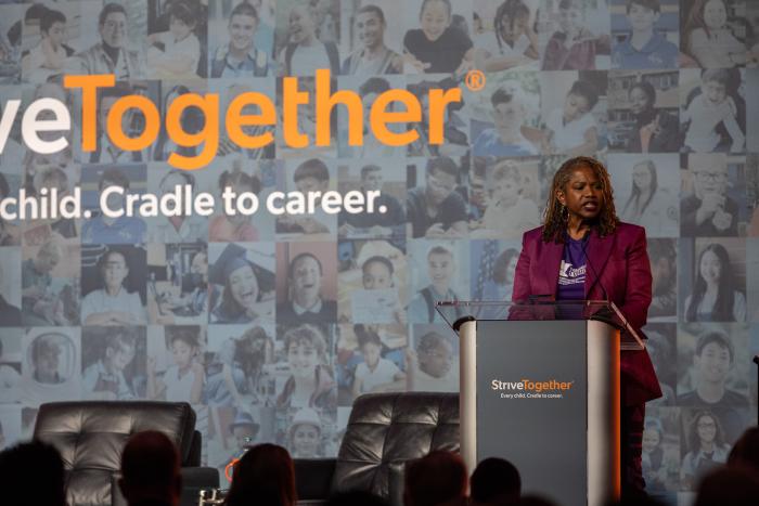 A Black woman stands at a podium wearing a purple blazer.