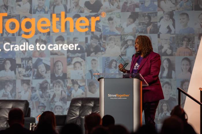 A Black woman stands at a podium wearing a purple blazer.