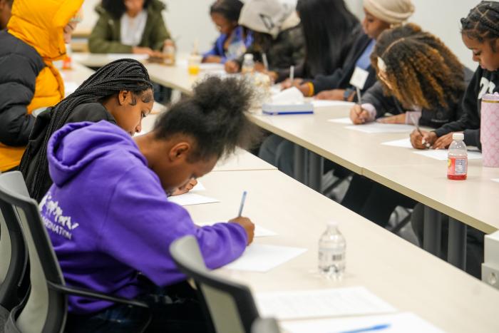 A young Black woman in a purple sweatshirt is writing on a piece of paper while sitting at a table.