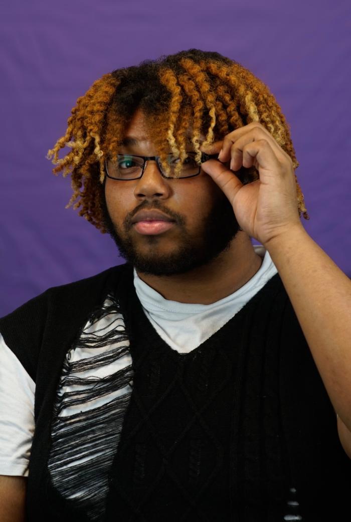 A young Black man in front of a purple background holds his glasses