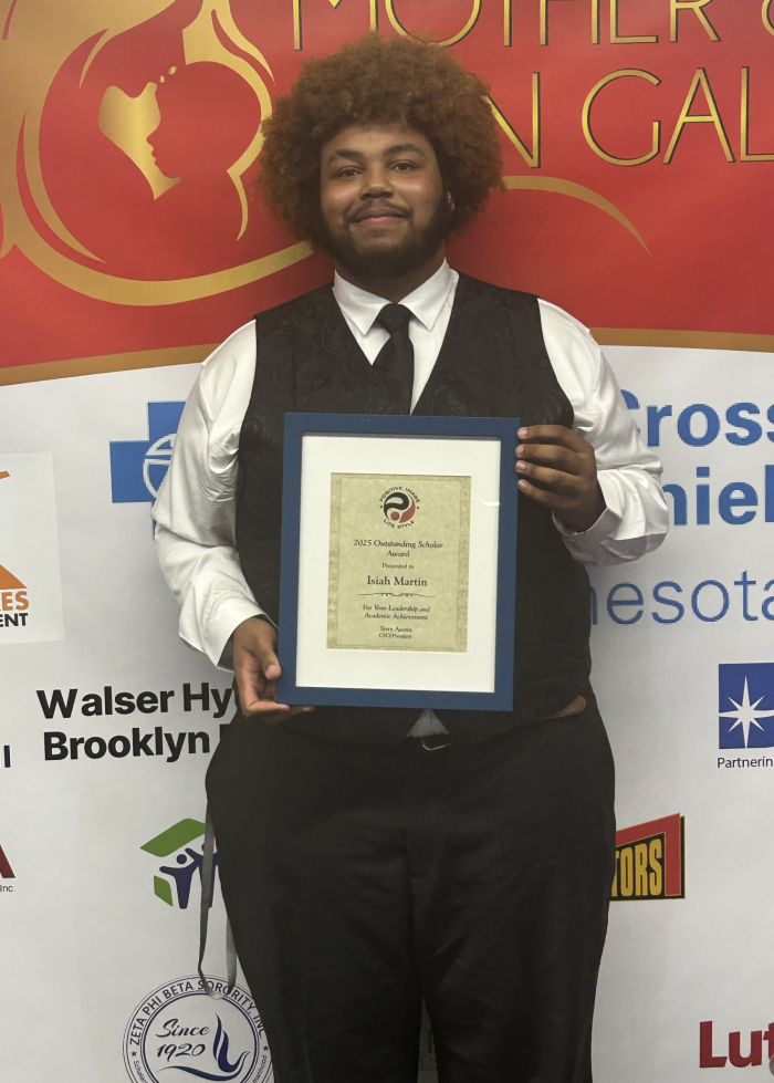 A young Black man in a white button down and black vest holds a certificate in front of him.