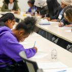A young Black woman in a purple sweatshirt is writing on a piece of paper while sitting at a table.